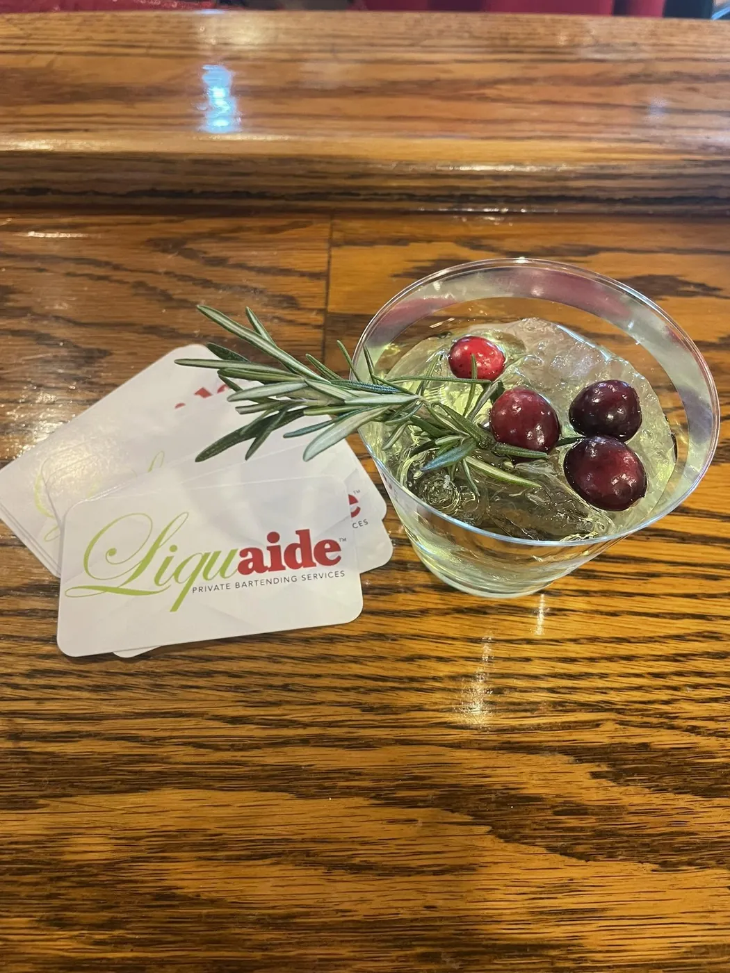 Signature cocktail garnished with rosemary and fresh cranberries beside Liquaide business cards